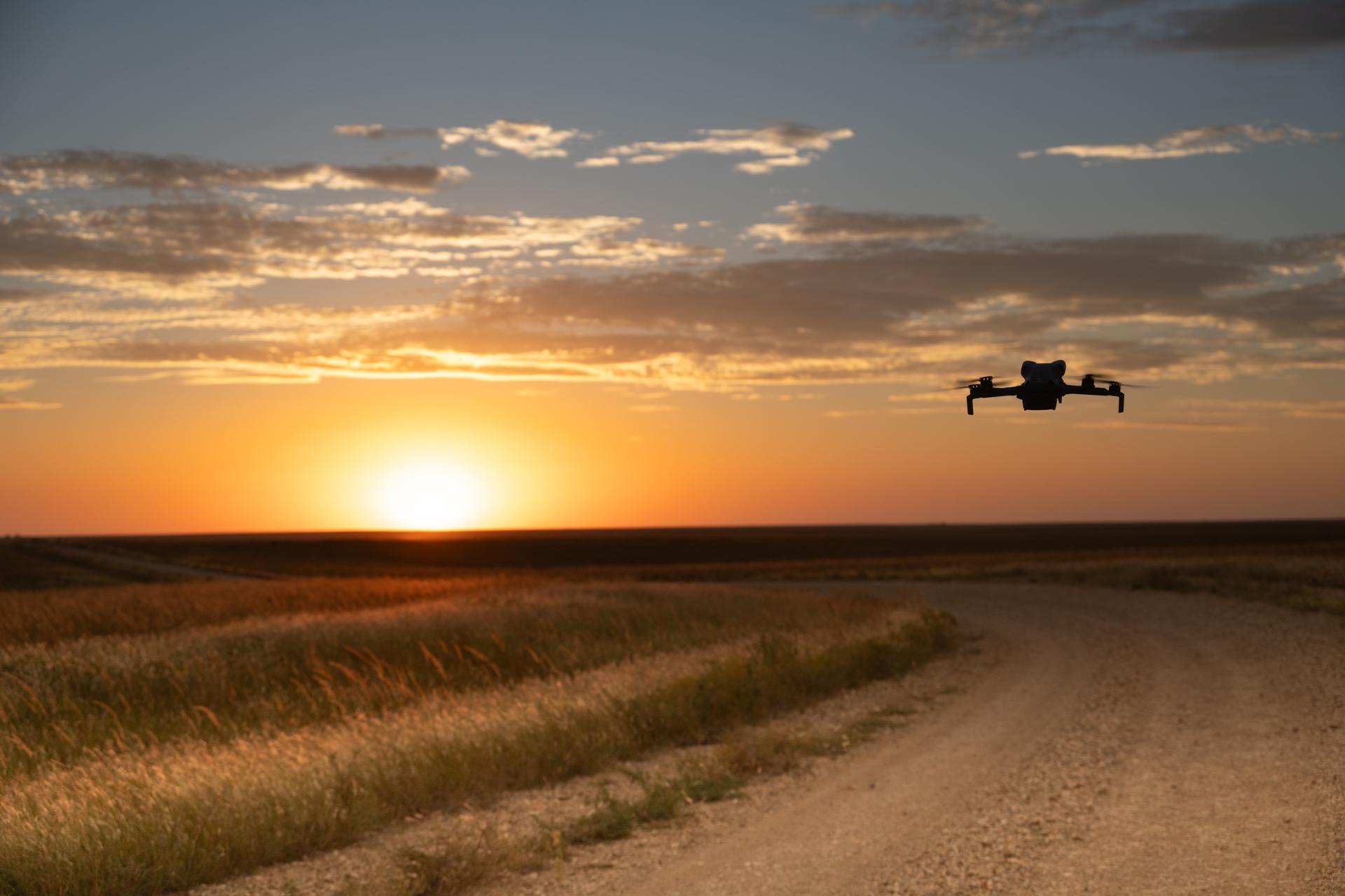 A small drone hovering over a gravel road through prairie grass at sunset
