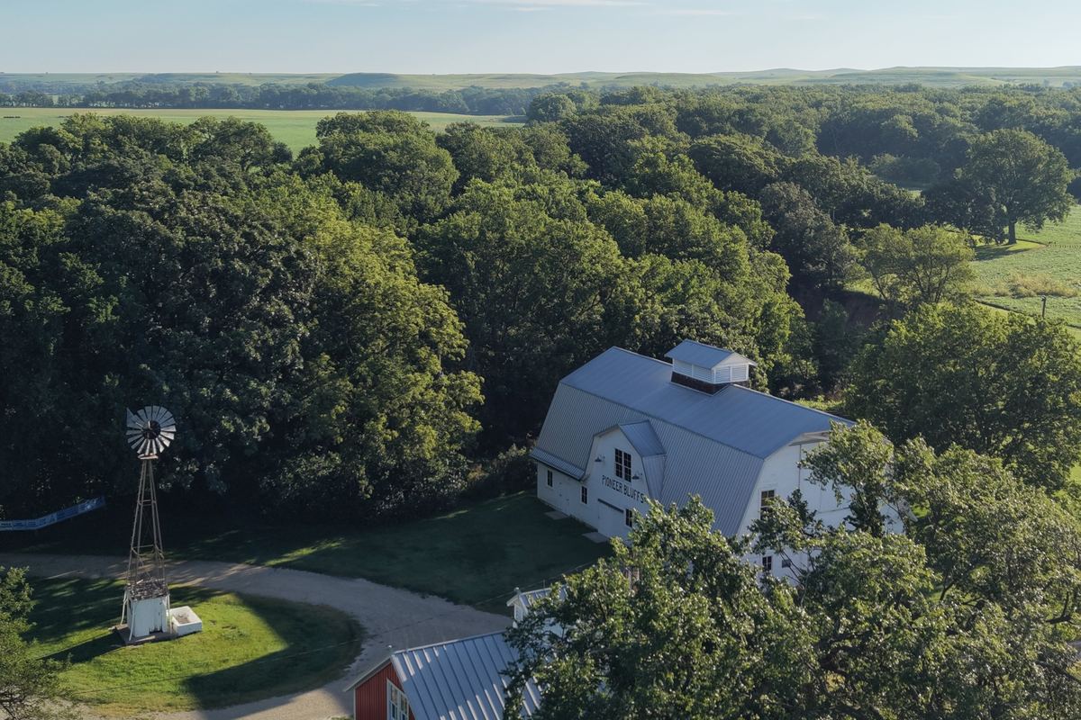 Aerial view of a white gambrel barn, red outbuilding, and windmill surrounded by trees in the Flint Hills